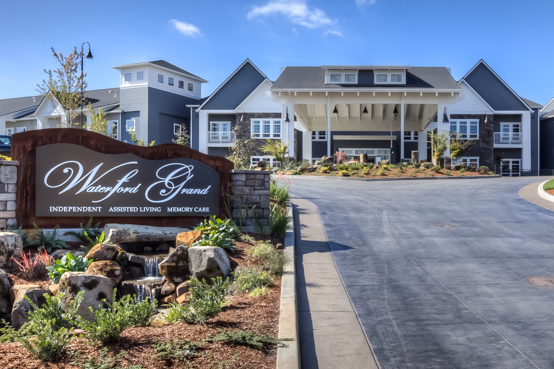 Entrance to the Waterford Grand senior living facility with a landscaped sign, small rock waterfall, and the building's covered driveway.