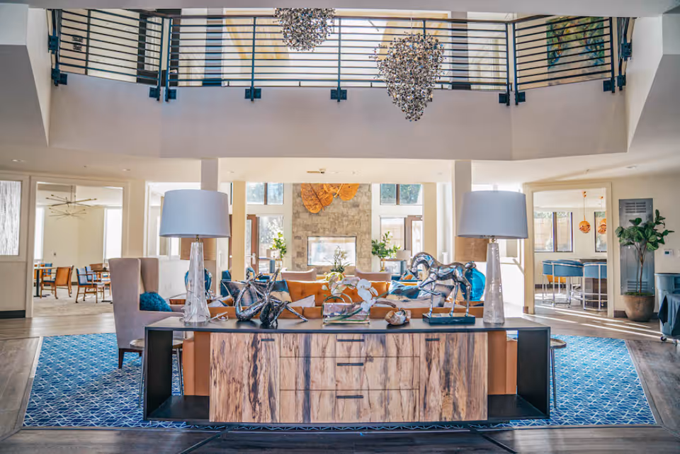 Spacious two-story common lounge with a decorative console table and lamps in the foreground, seating and a fireplace beyond, and a chandelier hanging from the upper level.