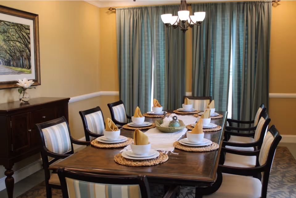 A formal dining room with a wooden table set for eight people. Each place setting includes a white bowl, plate, and a yellow folded napkin on a woven placemat. The chairs have striped upholstery. There is a sideboard with a flower vase and a framed picture on the wall. Blue curtains cover the windows, and a chandelier with six lights hangs above the table.