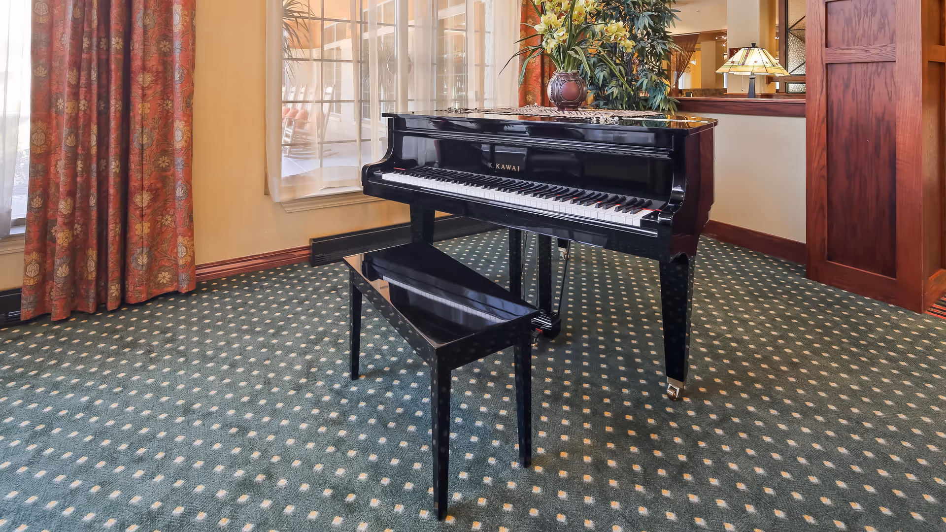 A black grand piano with a matching bench is placed on a green carpet with a white dotted pattern in a well-lit room. Behind the piano, there is a large window with sheer white curtains and red patterned drapes. To the right, there is a wooden panel and a table lamp with a stained glass shade, along with a large potted plant on a wooden surface.