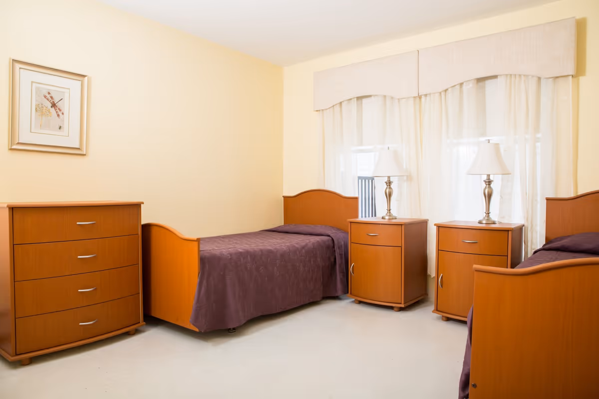 A simple, clean bedroom with two single wooden beds covered in purple bedspreads. Between the beds are two wooden nightstands, each with a white lamp. There is a wooden dresser with four drawers on the left side of the room and a framed picture hanging above it. Light-colored curtains cover the window behind the beds.