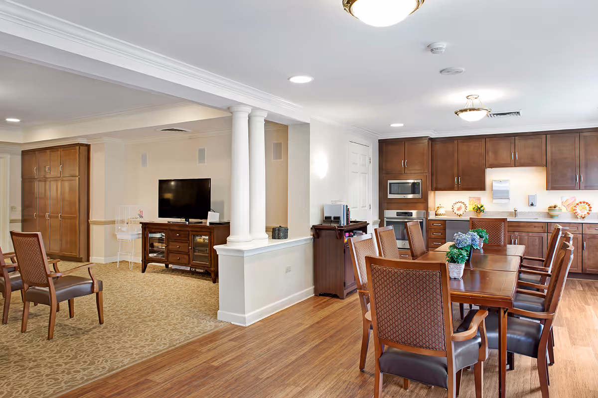 Open communal interior showing a dining table with chairs, kitchen cabinets and appliances, a TV area, and decorative columns.