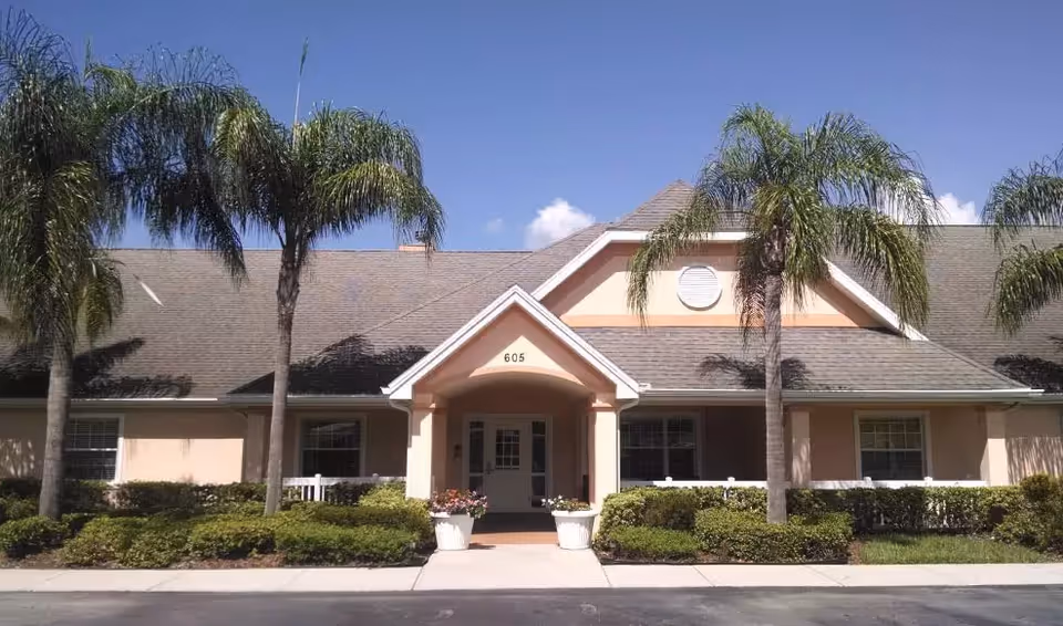 Front exterior view of a single-story building with a peaked roof, beige walls, and white trim. The entrance has a small covered porch with the number 605 above the door. There are palm trees and well-maintained bushes in front of the building under a clear blue sky.