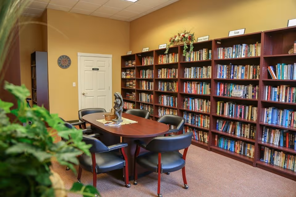 A cozy library room with wooden bookshelves filled with books organized by categories such as Poetry/Songbooks and Fiction. There is a wooden table in the center surrounded by six black chairs with red legs. A decorative statue and a basket are placed on the table. The walls are painted a warm yellow color, and a clock hangs near a white door labeled 'Employees Only'. A green plant is partially visible in the foreground.