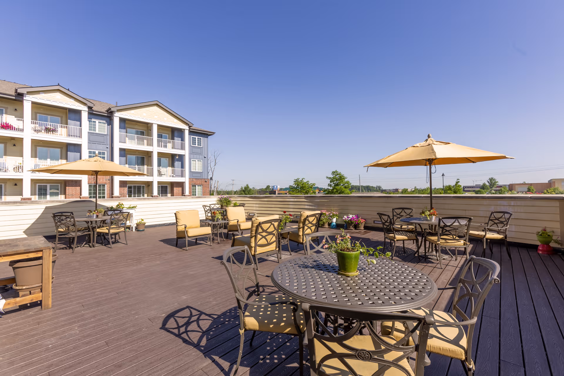 Outdoor patio area with multiple metal tables and chairs, some with beige cushions and umbrellas. Potted plants and flowers decorate the space. A multi-story residential building is visible in the background under a clear blue sky.