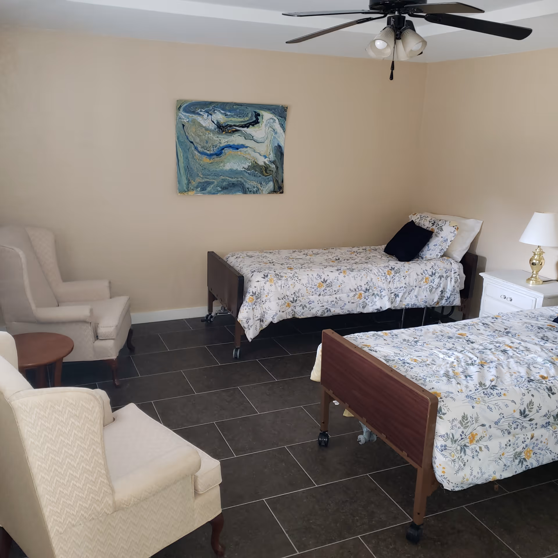 A shared bedroom with two twin hospital-style beds with floral bedspreads, armchairs, a nightstand with lamp, and wall art.