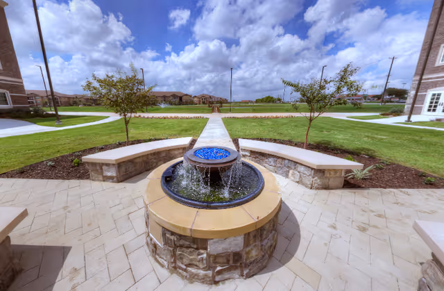 Circular stone fountain with blue glass beads in a paved courtyard flanked by curved benches and grassy lawns between brick buildings under a partly cloudy sky.