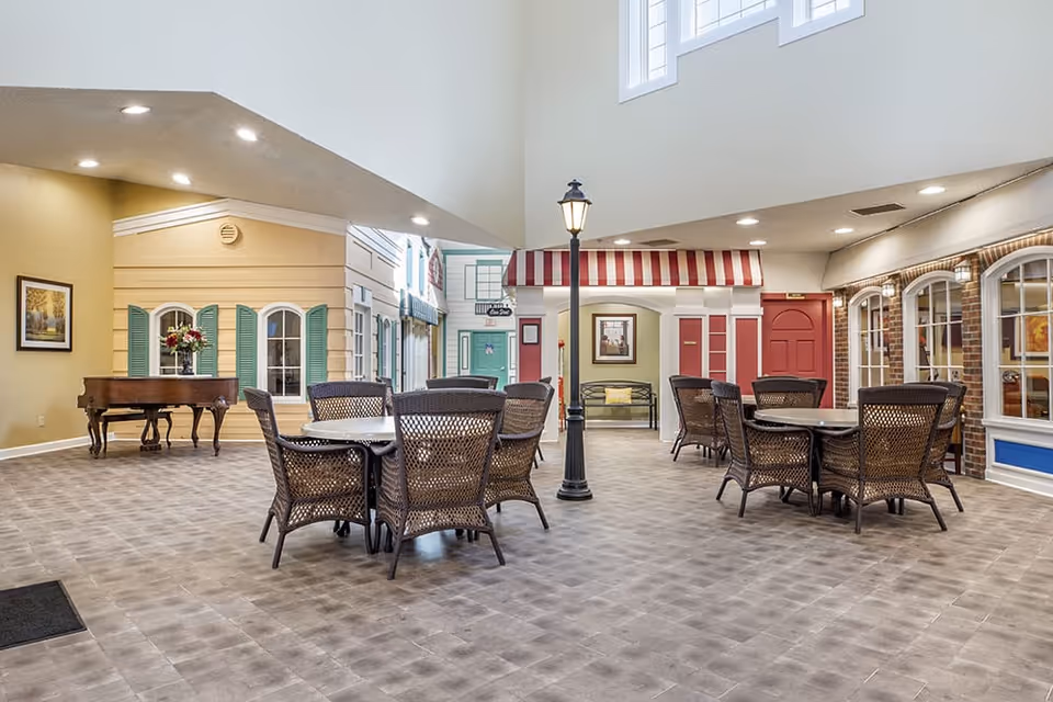 Indoor common area with multiple round tables and wicker chairs arranged around a central lamp post. The space features high ceilings with recessed lighting, decorative facades resembling storefronts with windows and doors, and a grand piano with a flower arrangement on top. The floor is tiled with a gray pattern.