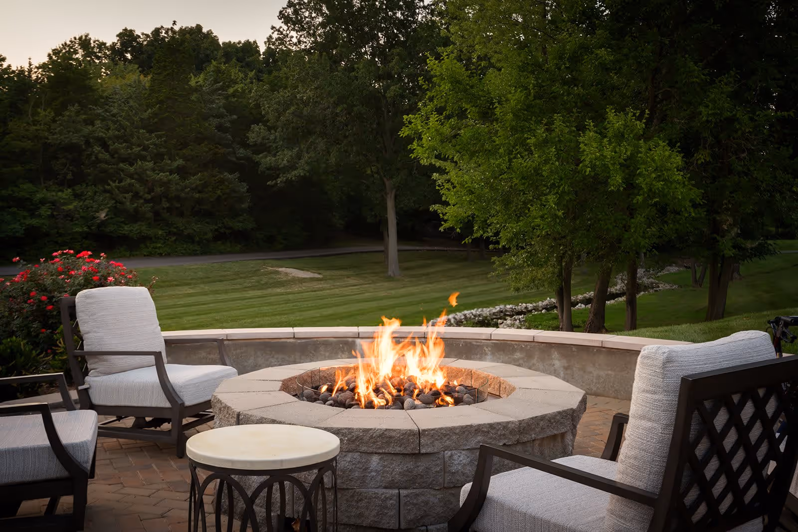 Outdoor patio area with cushioned chairs arranged around a stone fire pit with flames, surrounded by green trees and a well-maintained lawn.
