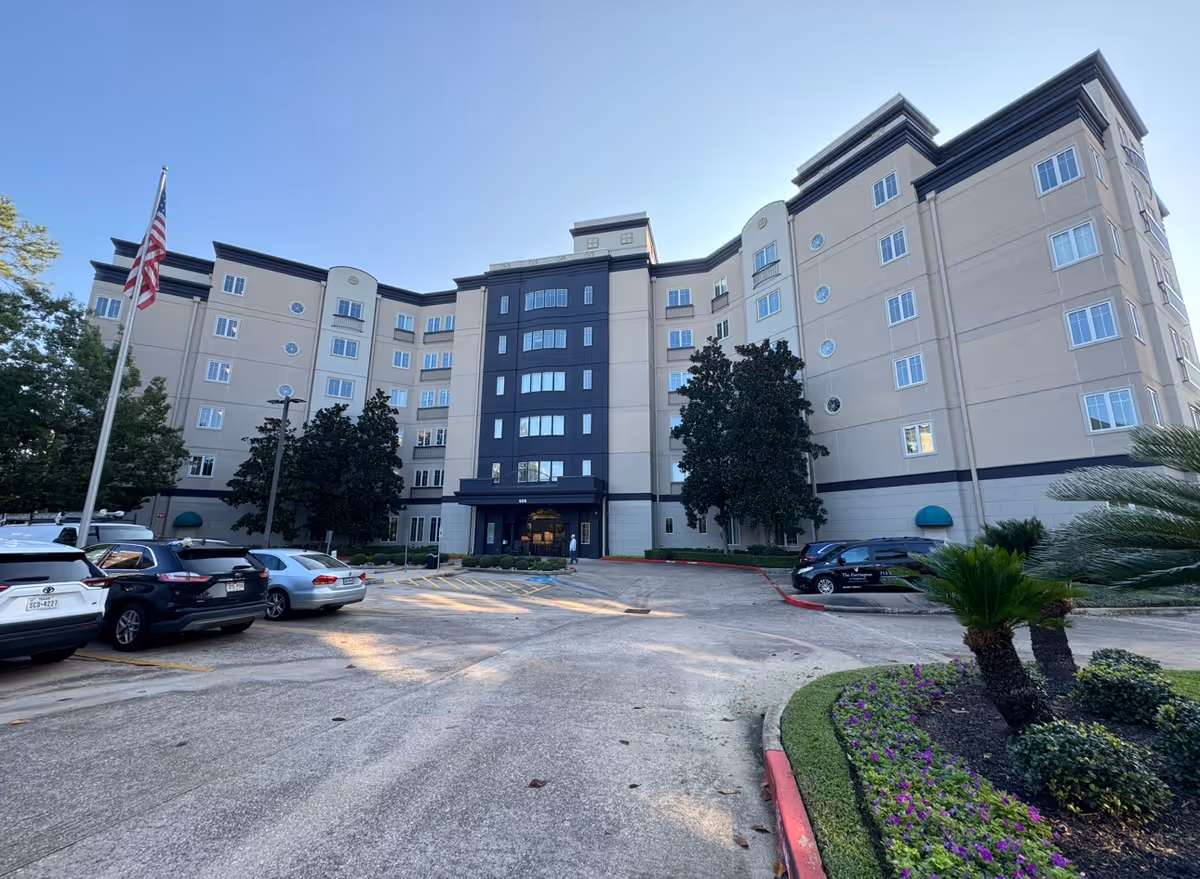 Exterior view of a multi-story senior living facility building with several windows, an entrance canopy, a parking lot with cars, an American flag on a flagpole, and landscaped greenery including trees and bushes under a clear blue sky.