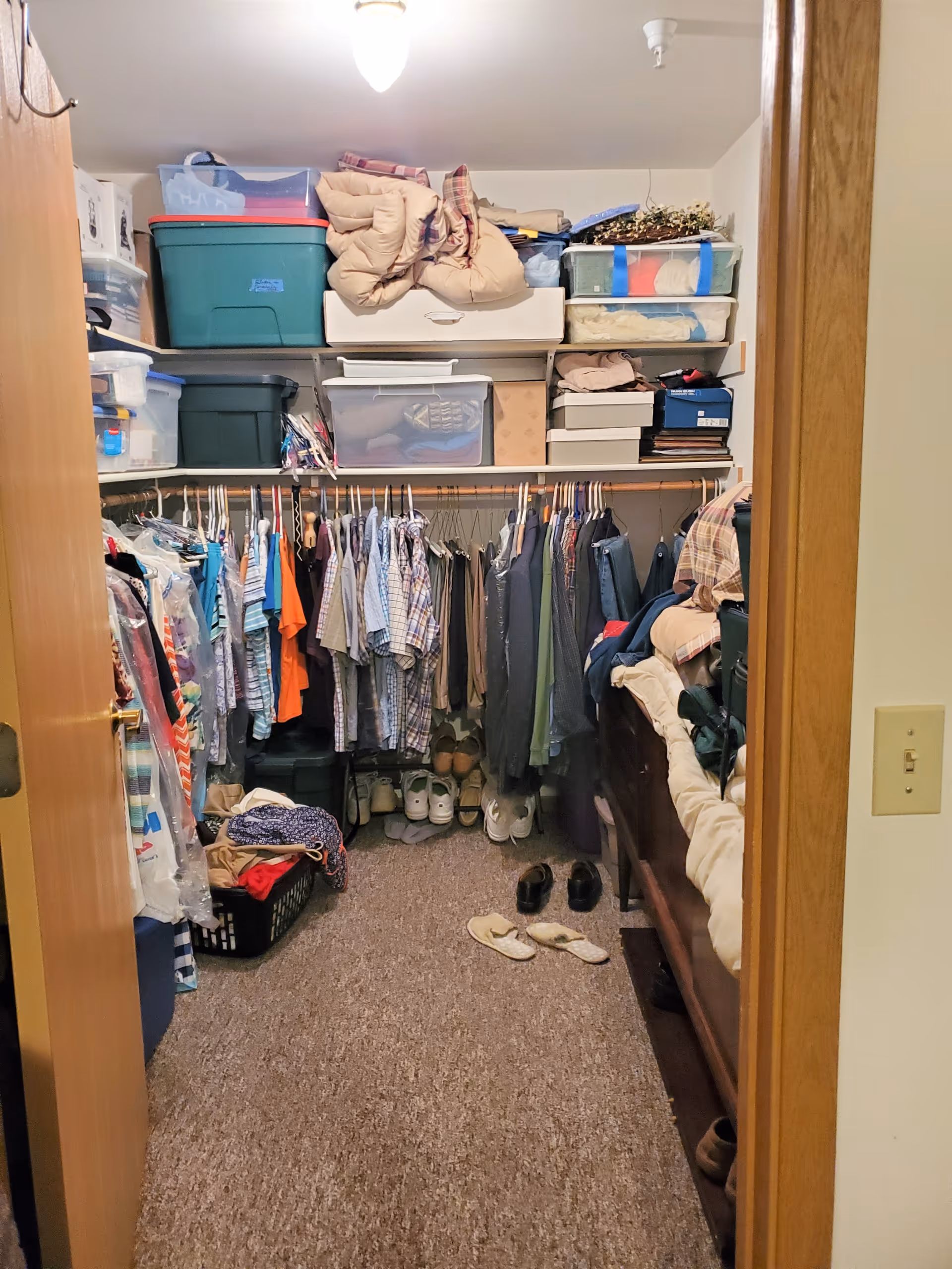 View into a walk-in closet with clothes hanging on rods along the left and back walls, shelves above holding storage bins and folded items, shoes lined up on the floor, and a bed partially visible on the right side.