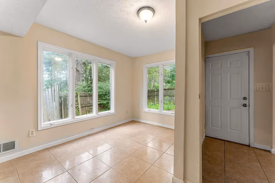 Bright empty room with large windows overlooking a fenced yard and a closed white entry door.