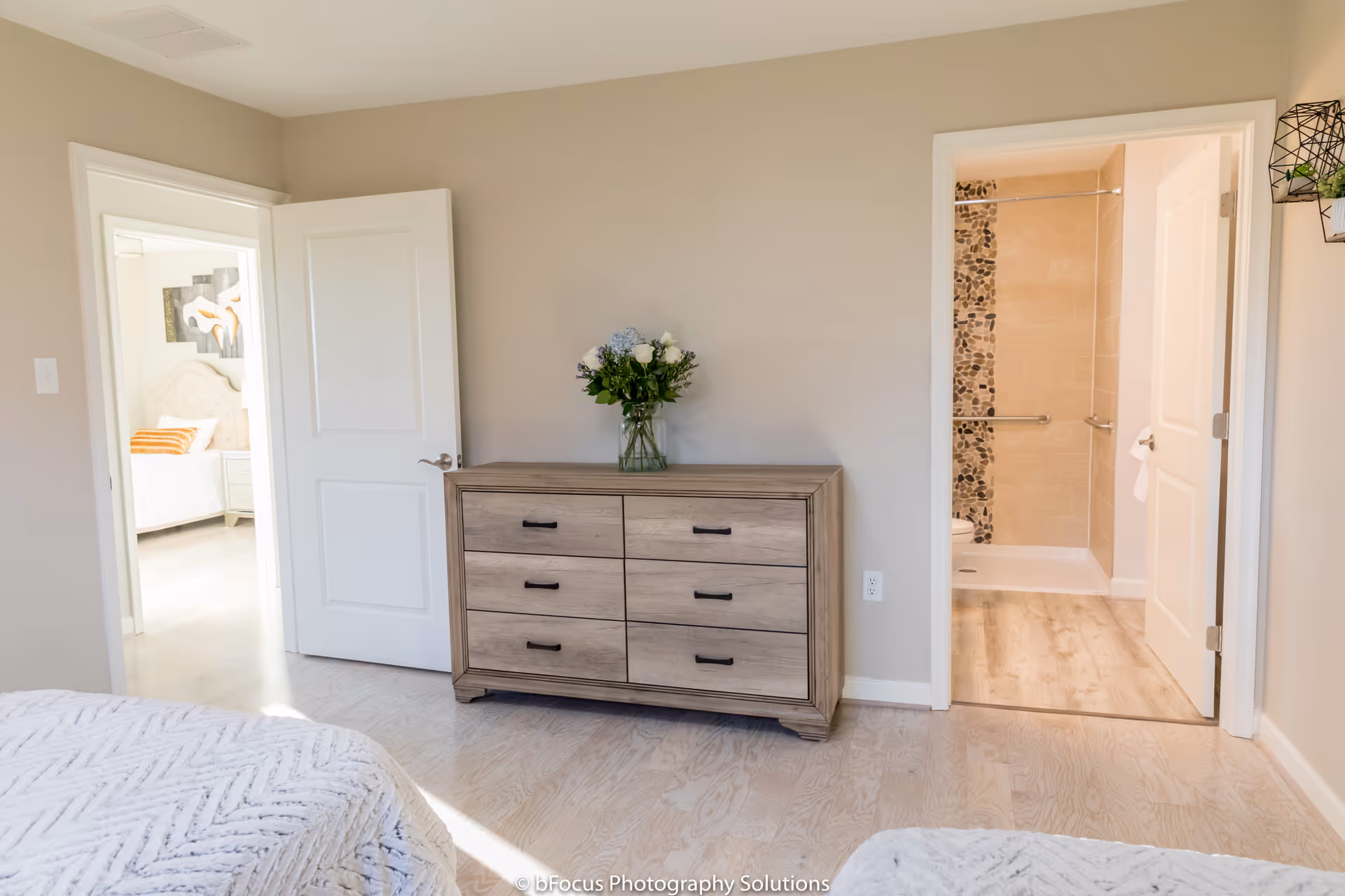 A bedroom with light-colored walls and wood flooring. There is a wooden dresser with six drawers against the wall, topped with a vase of flowers. Two open doors are visible: one leading to another bedroom with a bed and artwork on the wall, and the other leading to a bathroom with a walk-in shower featuring pebble tile accents and grab bars.