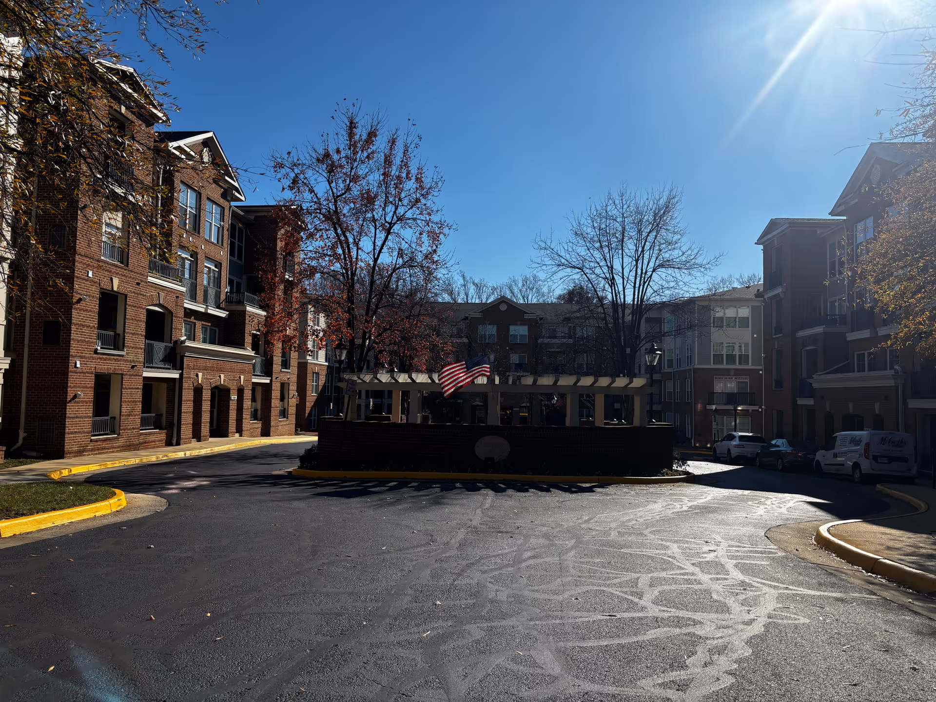 Entrance driveway and courtyard of a multi-story brick senior living complex with an American flag under a clear blue sky.