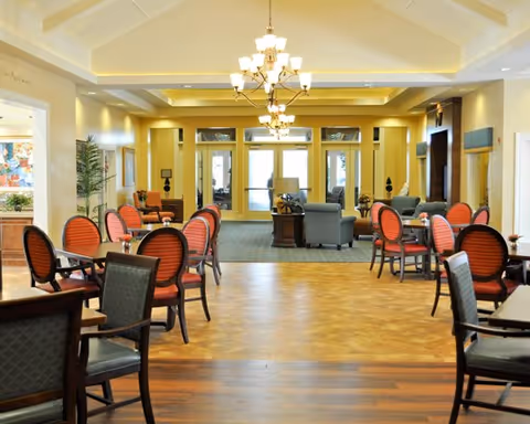 A spacious and well-lit common area in a senior living facility with multiple tables and chairs arranged for seating. The room features a high ceiling with chandeliers, a mix of carpet and wood flooring, and comfortable lounge chairs near the back by large glass doors leading outside. The walls are painted in warm tones, and there are decorative plants and lamps placed around the room.
