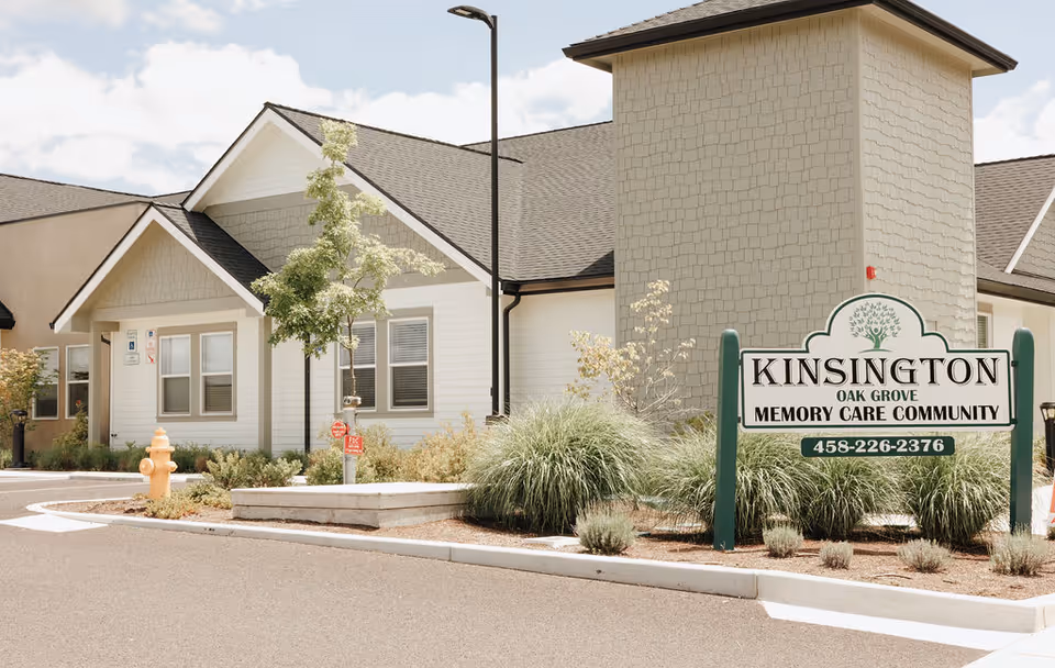 Exterior view of Kinsington Oak Grove Memory Care Community building with a sign displaying the facility name and phone number. The building has a light-colored facade with a gabled roof, surrounded by landscaping including bushes and a small tree. A fire hydrant and a street lamp are also visible near the entrance.