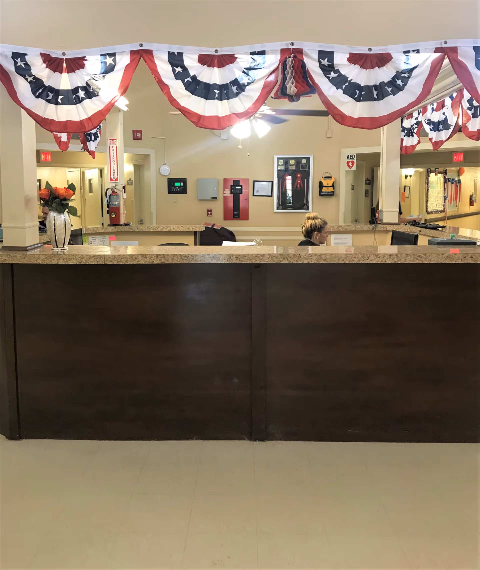 Reception desk area in a senior living facility decorated with red, white, and blue patriotic bunting. A woman is seated behind the desk, and there are various safety and informational signs on the wall behind her, including a fire extinguisher and an AED device.