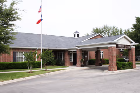 Exterior view of a single-story brick building with a covered entrance supported by brick columns. There are two flagpoles with the American and Texas flags in front of the building, surrounded by green shrubs and trees.