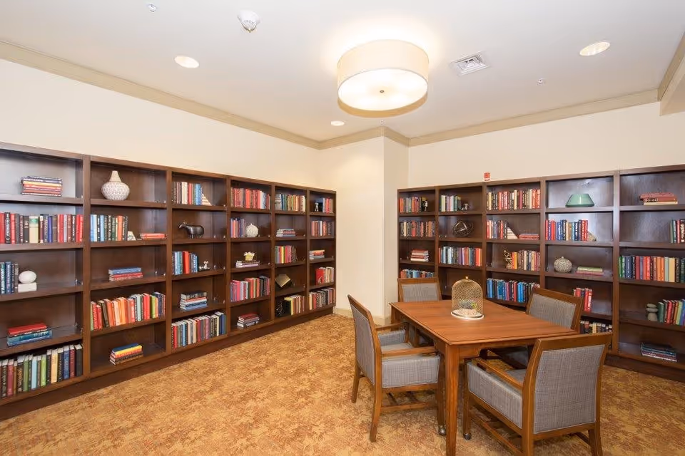 Well-lit library room with wooden bookshelves lining the walls and a central table surrounded by chairs.