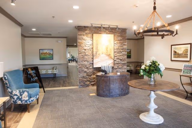 A welcoming interior lobby area of The Aspen of Brookhaven featuring a round wooden table with a vase of white flowers, a stone accent wall with a large illuminated painting, a blue upholstered chair with floral cushions, a small round reception desk, and framed artwork on the walls.