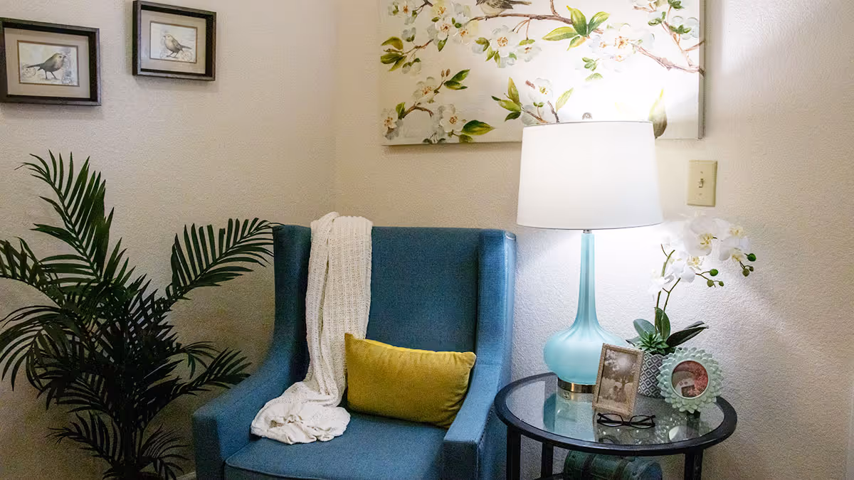 A cozy corner with a blue armchair draped with a white knitted throw and a yellow cushion. Next to the chair is a round glass-top side table holding a blue lamp, a small potted orchid, two framed photos, and a pair of glasses. On the wall above the chair is a floral painting and two small framed bird pictures. A green leafy plant is placed in the corner.