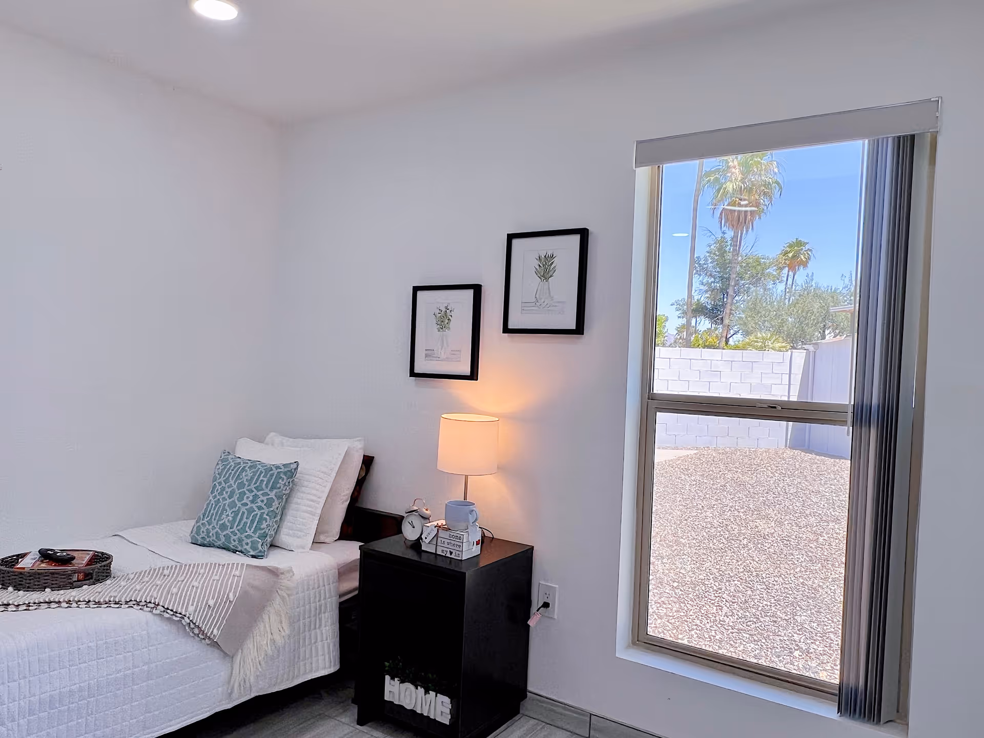 A small, neatly arranged bedroom with a single bed covered in white bedding and a decorative blue pillow. Next to the bed is a black nightstand with a lamp, an alarm clock, a small plant, and a cup. Two framed botanical prints hang on the white wall above the nightstand. A large window with vertical blinds shows an outdoor view of a gravel yard and palm trees under a clear blue sky.