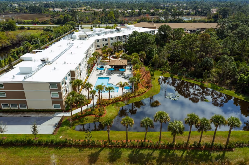 Aerial view of a multi-story senior living building with a central pool area and an adjacent pond surrounded by palm trees.