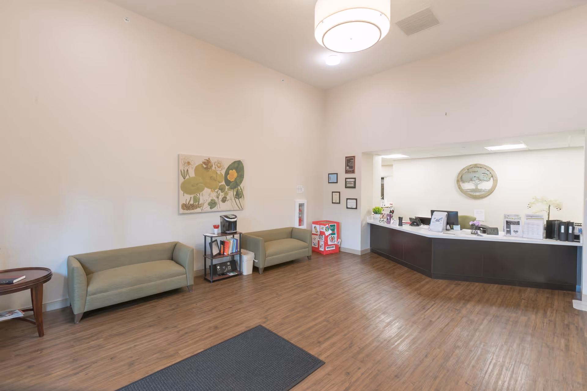 Reception area of a senior living facility with two green sofas, a small table with a coffee maker and books, a dark wood reception desk with office supplies and decorations, light wood flooring, and a large round ceiling light fixture.