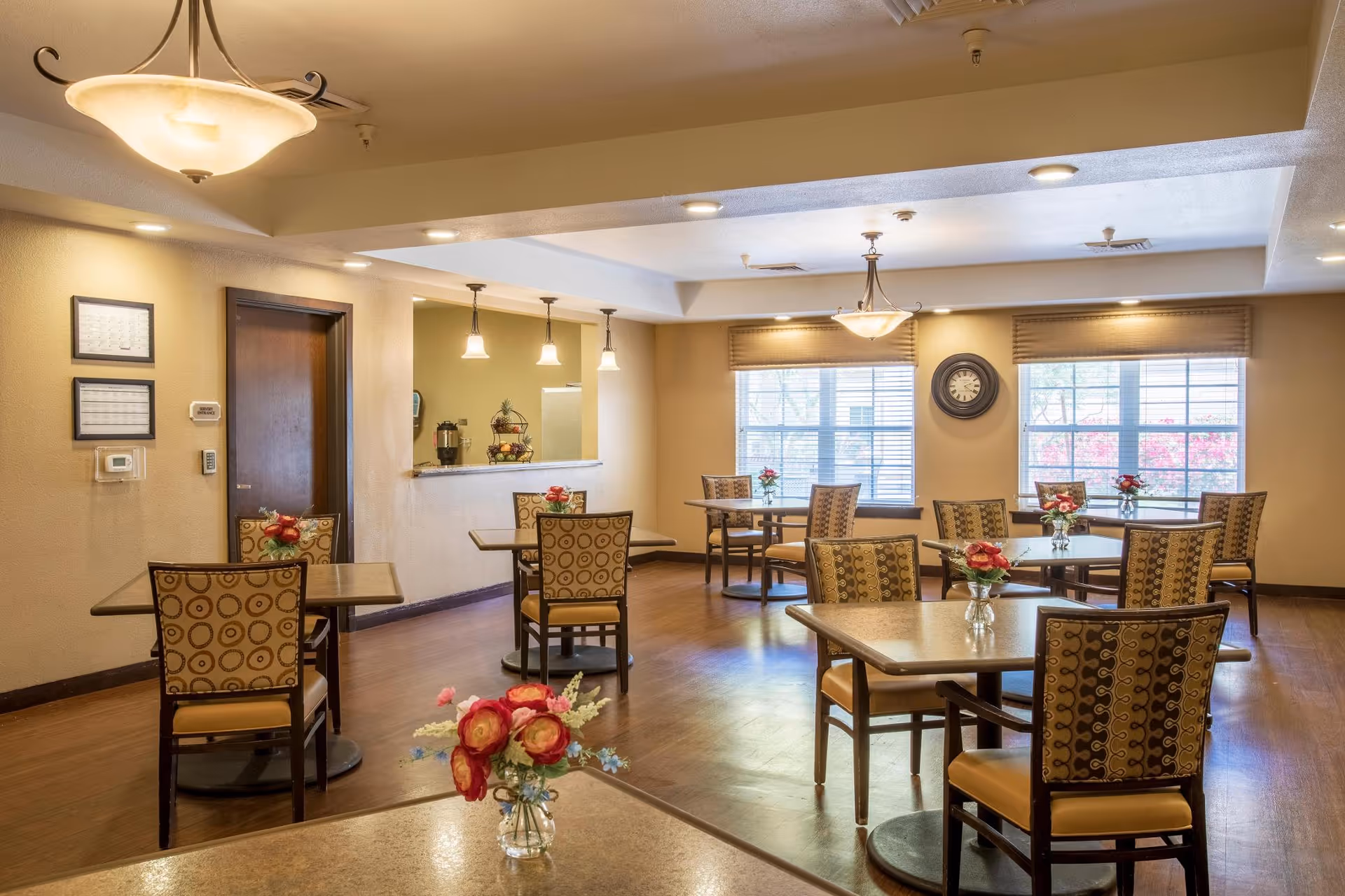 A well-lit dining room with multiple square tables and patterned chairs. Each table has a small vase with red and pink flowers. There are large windows with blinds letting in natural light, a wall clock between the windows, and ceiling lights providing additional illumination. The room has warm beige walls and wooden flooring.