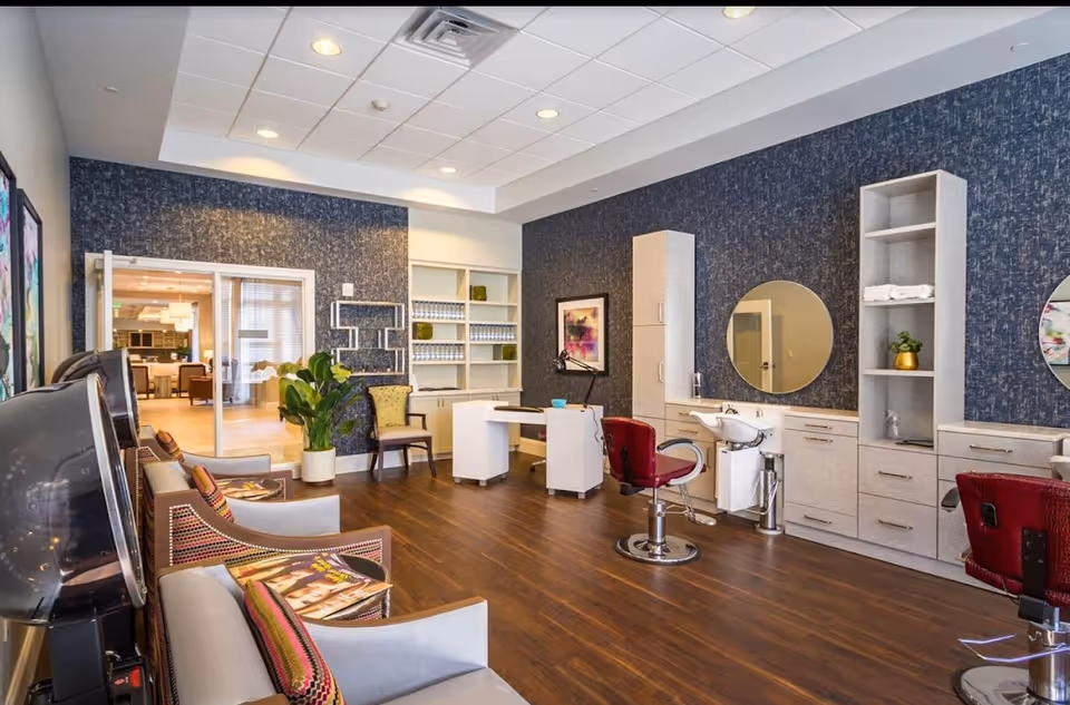 Bright salon-style interior with styling chairs, wash sinks, shelving and a seating area in a senior living facility.