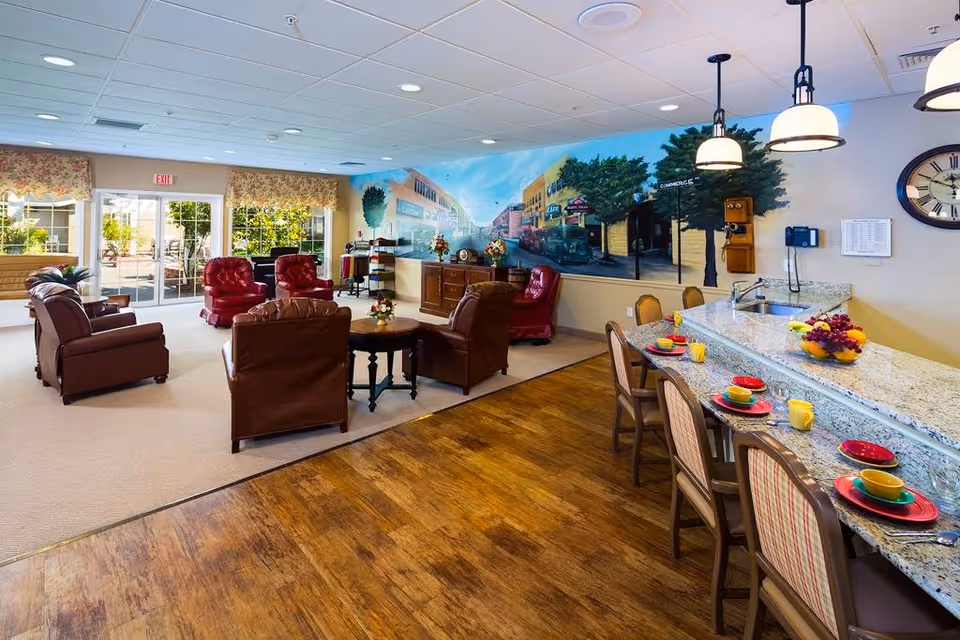 A spacious and well-lit common area in a senior living facility featuring a seating area with leather armchairs and a round wooden table. The room has a large mural on one wall depicting a vintage street scene. Adjacent to the seating area is a granite countertop with several chairs set with colorful dishes and cups. Large windows and glass doors provide natural light and a view of an outdoor patio area.