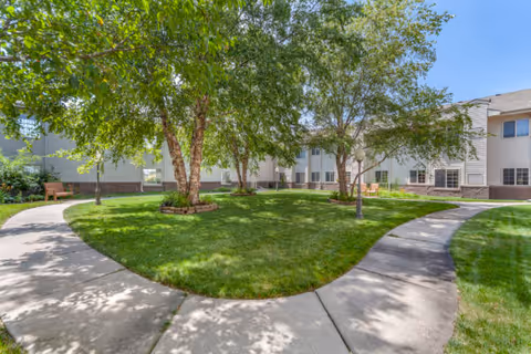 A landscaped outdoor courtyard area with a circular concrete pathway surrounding a grassy area with several trees. The courtyard is bordered by a two-story building with multiple windows. There is a bench visible on the left side under the trees.