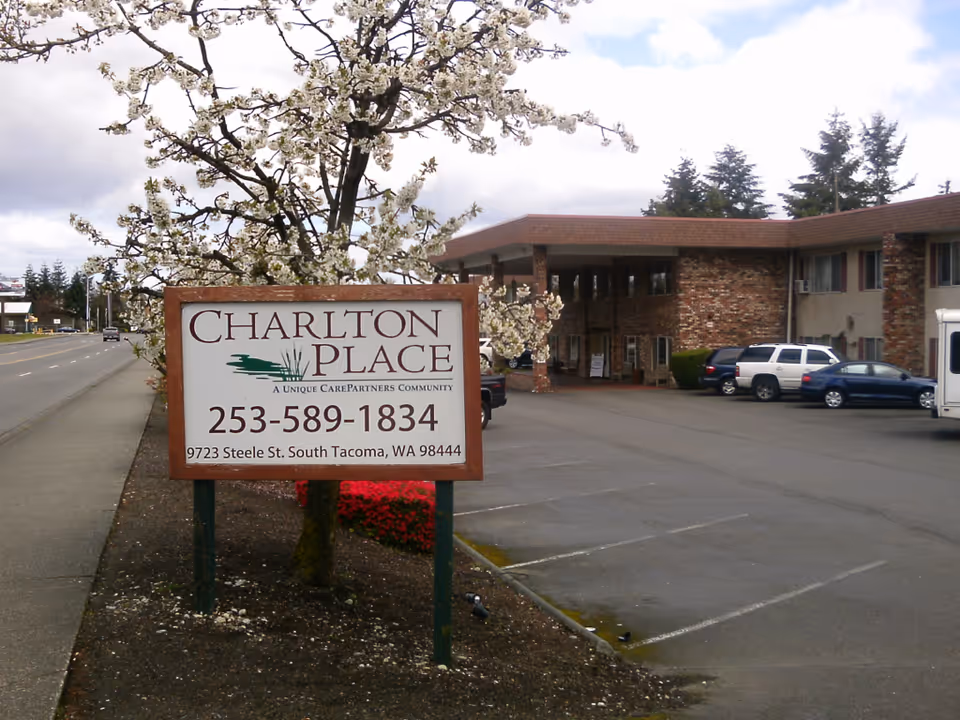 Exterior view of Charlton Place senior living facility with a sign displaying the name, phone number, and address. The building is a two-story structure with brick and beige walls, a covered entrance, and a parking lot with several parked cars. A blossoming tree is next to the sign, and a sidewalk runs alongside the street.