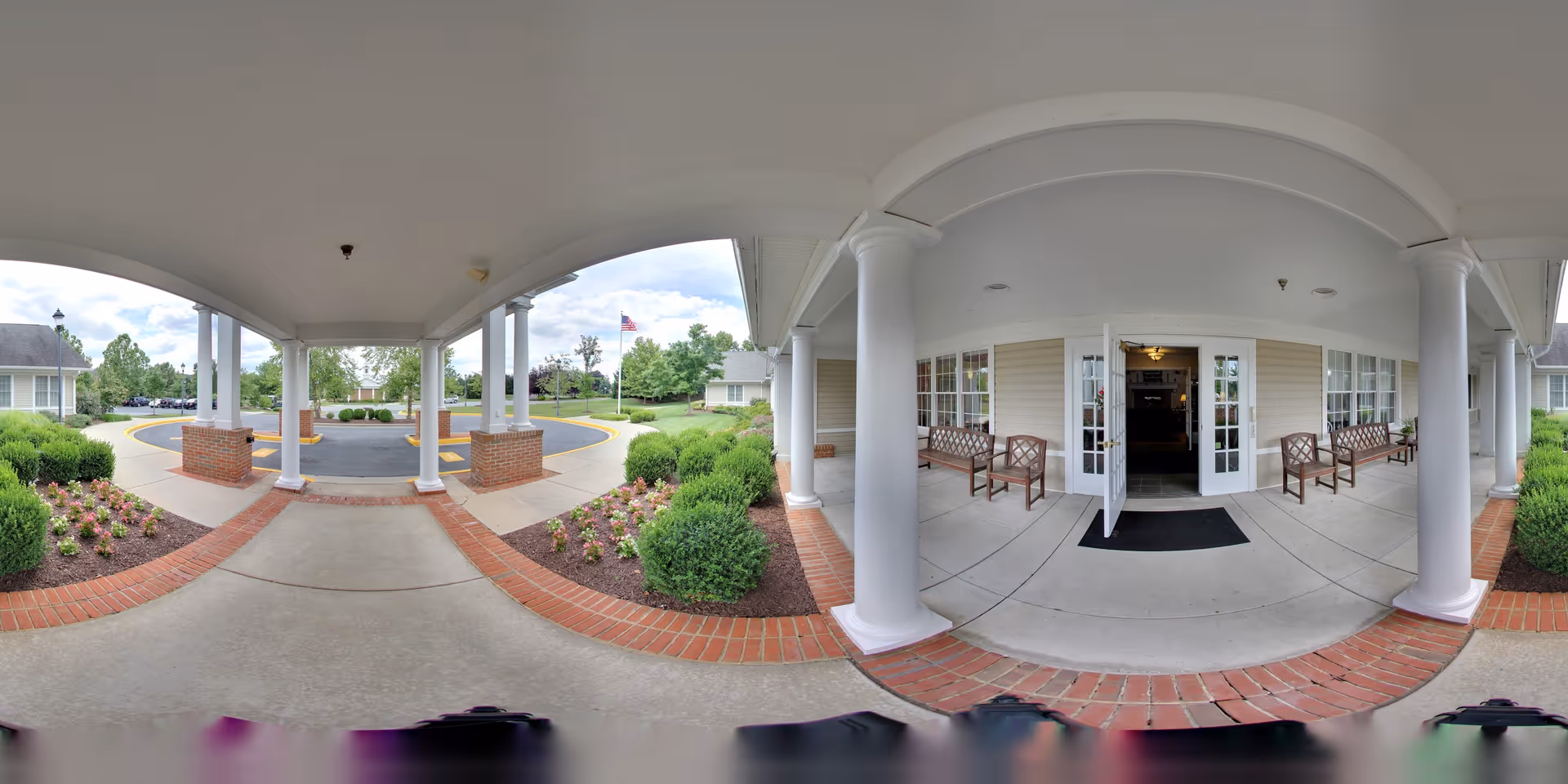 Covered entrance area of Heart Fields Assisted Living At Frederick with white columns, brick accents, and benches on either side of the open double doors leading inside. There are landscaped flower beds with bushes and flowers on both sides of the walkway, and an American flag is visible in the background.