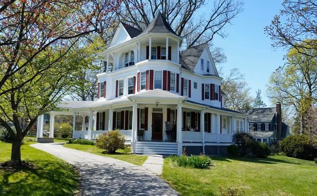 Large white Victorian-style house with a wraparound porch and red shutters set on a green lawn with trees.
