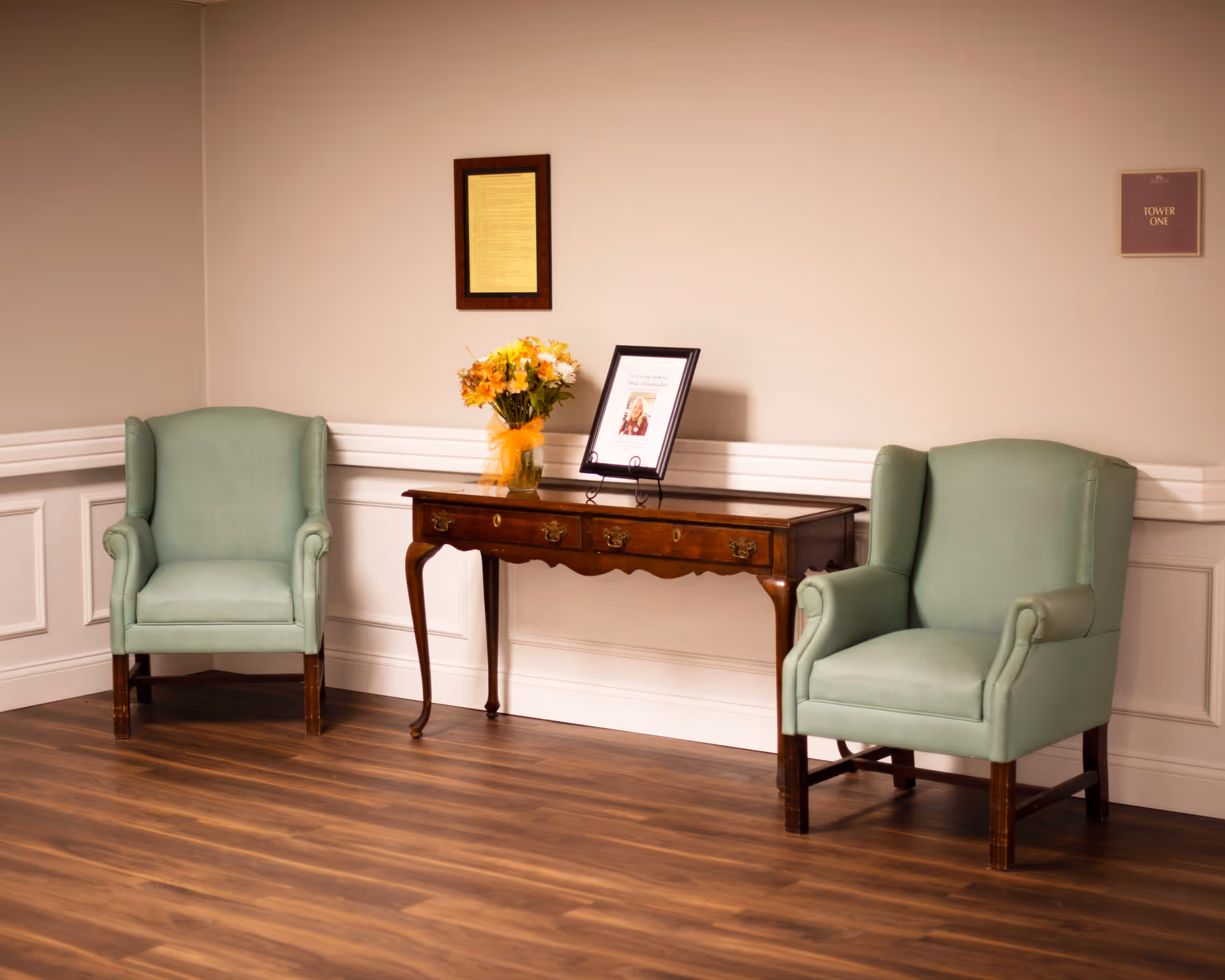 A cozy seating area with two light green upholstered armchairs placed on either side of a wooden console table. The table holds a vase of yellow flowers and a framed photo or document. The room has light beige walls with white wainscoting and wood flooring. A framed document and a small sign labeled 'Tower One' are mounted on the wall.