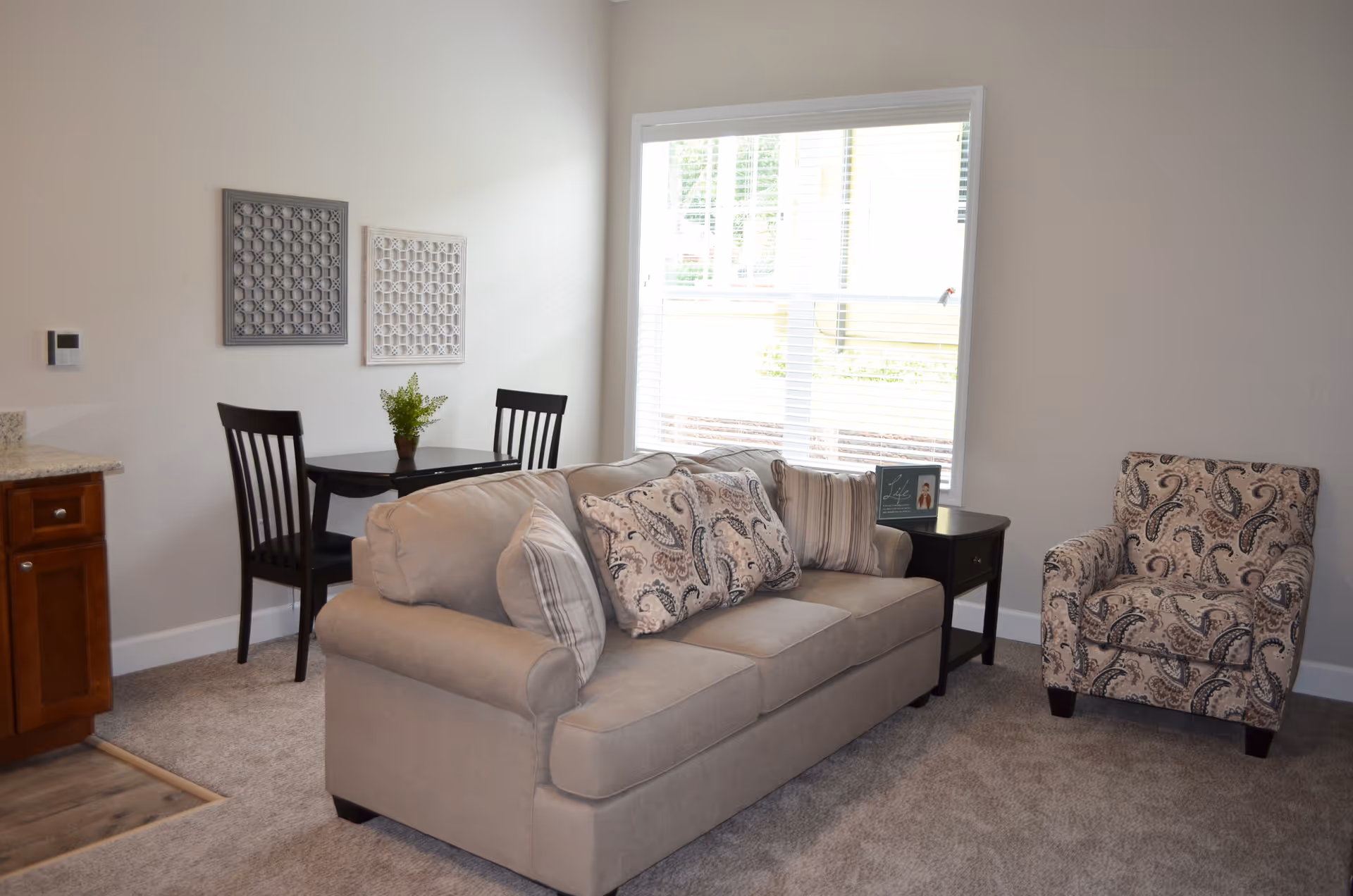 Sunny living area with a beige sofa, patterned armchair, small dining table and chairs beside a large window.