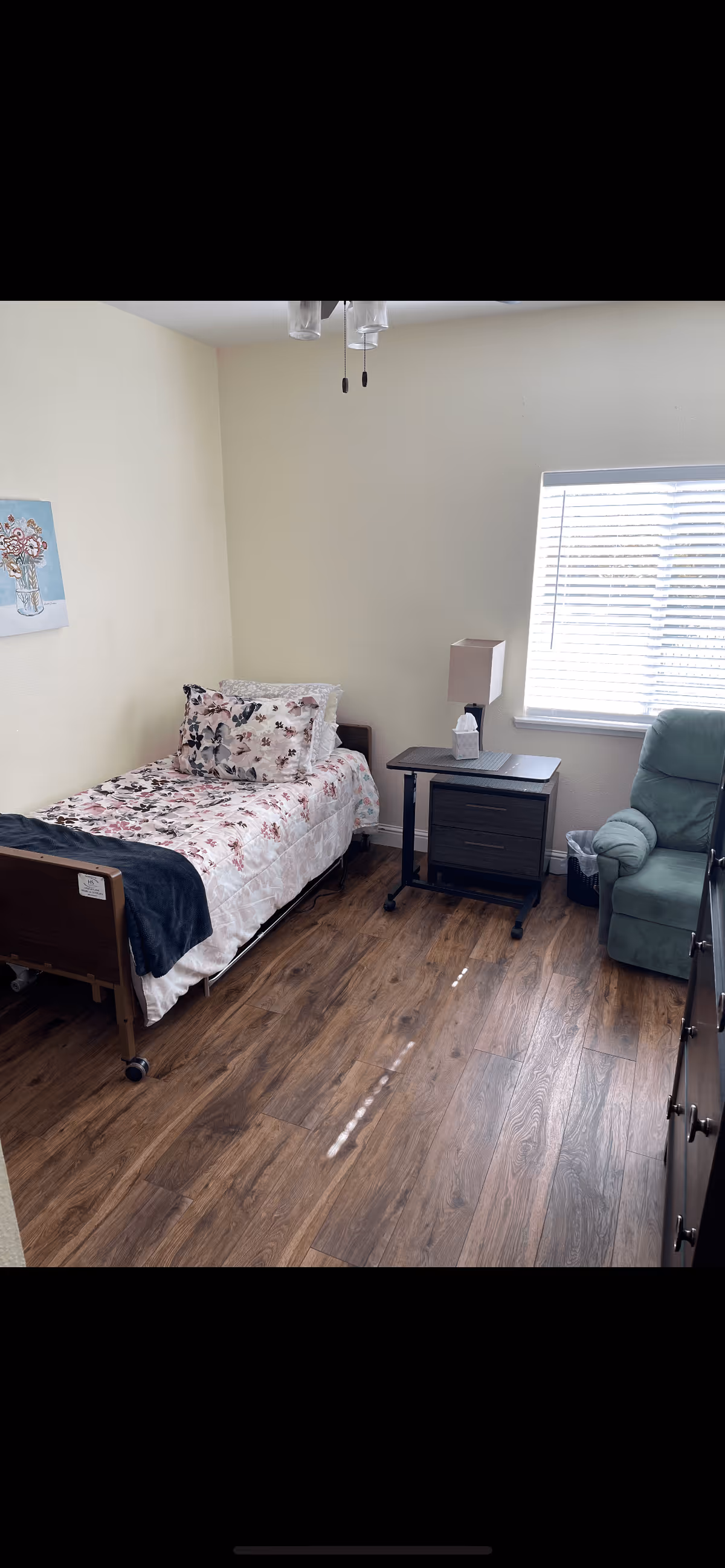 A small bedroom with a single bed covered in floral bedding, a bedside table with a lamp and tissue box, a green upholstered armchair, a window with blinds, and wooden flooring.