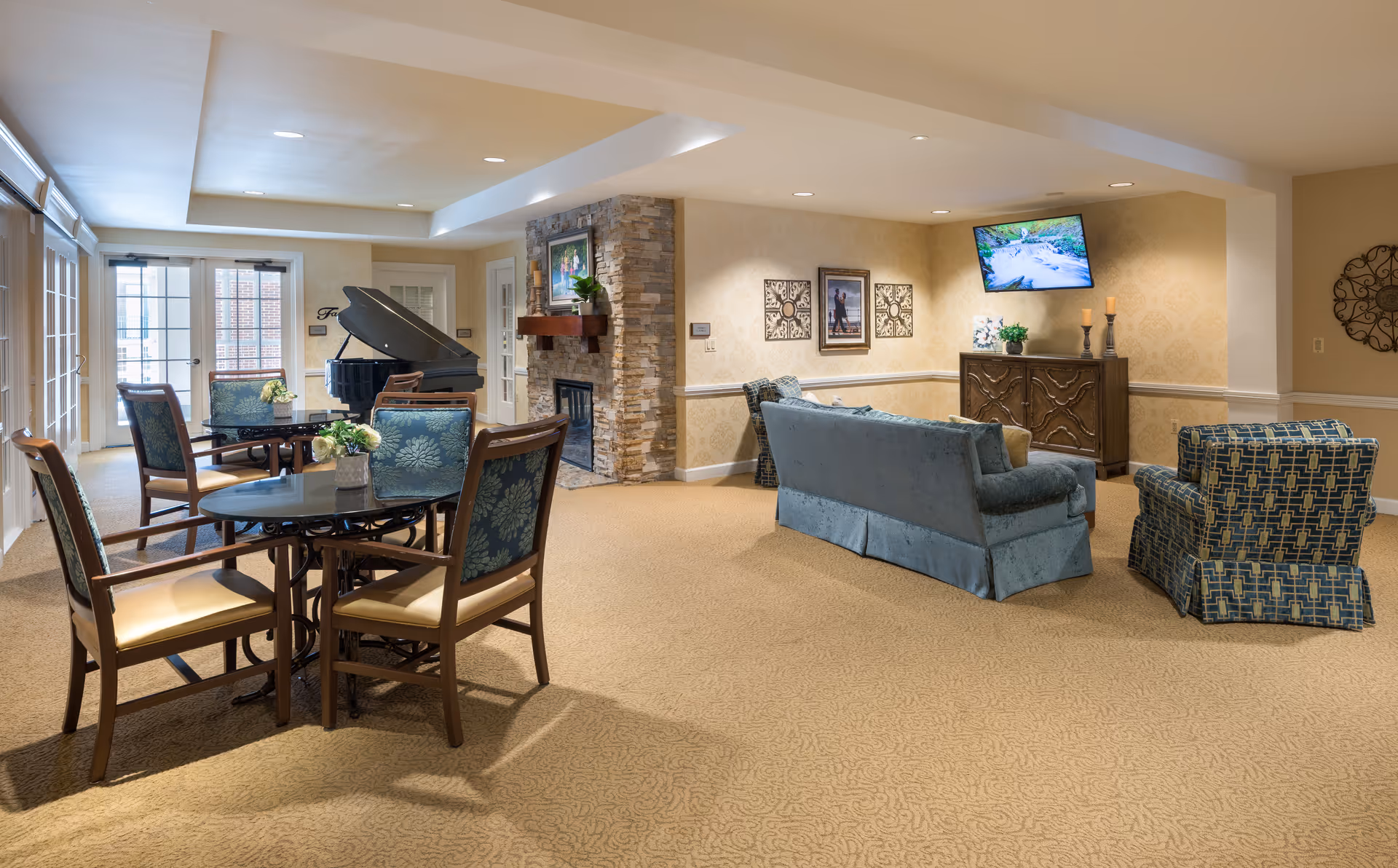 A spacious and well-lit common area in a senior living facility featuring a seating area with a blue sofa and patterned armchair, a wooden cabinet with a flat-screen TV mounted above it, a stone fireplace with a mantel, and a round table with four chairs. A grand piano is visible near the glass doors leading outside.