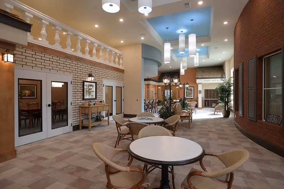 Interior view of a spacious common area in a senior living facility with round tables and wicker chairs, decorative plants, and a post office section in the background. The area features a mix of brick and painted walls, ceiling lights, and a cozy, inviting atmosphere.