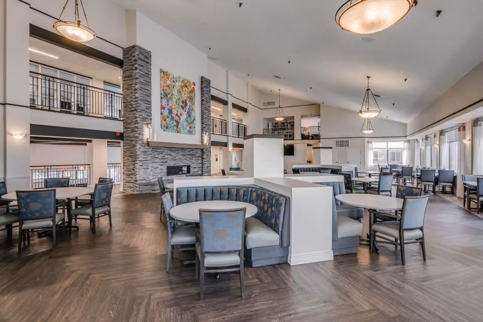 Spacious dining area in a retirement community with multiple round tables and chairs, a large stone fireplace with colorful artwork above it, high ceilings with hanging light fixtures, and large windows letting in natural light.