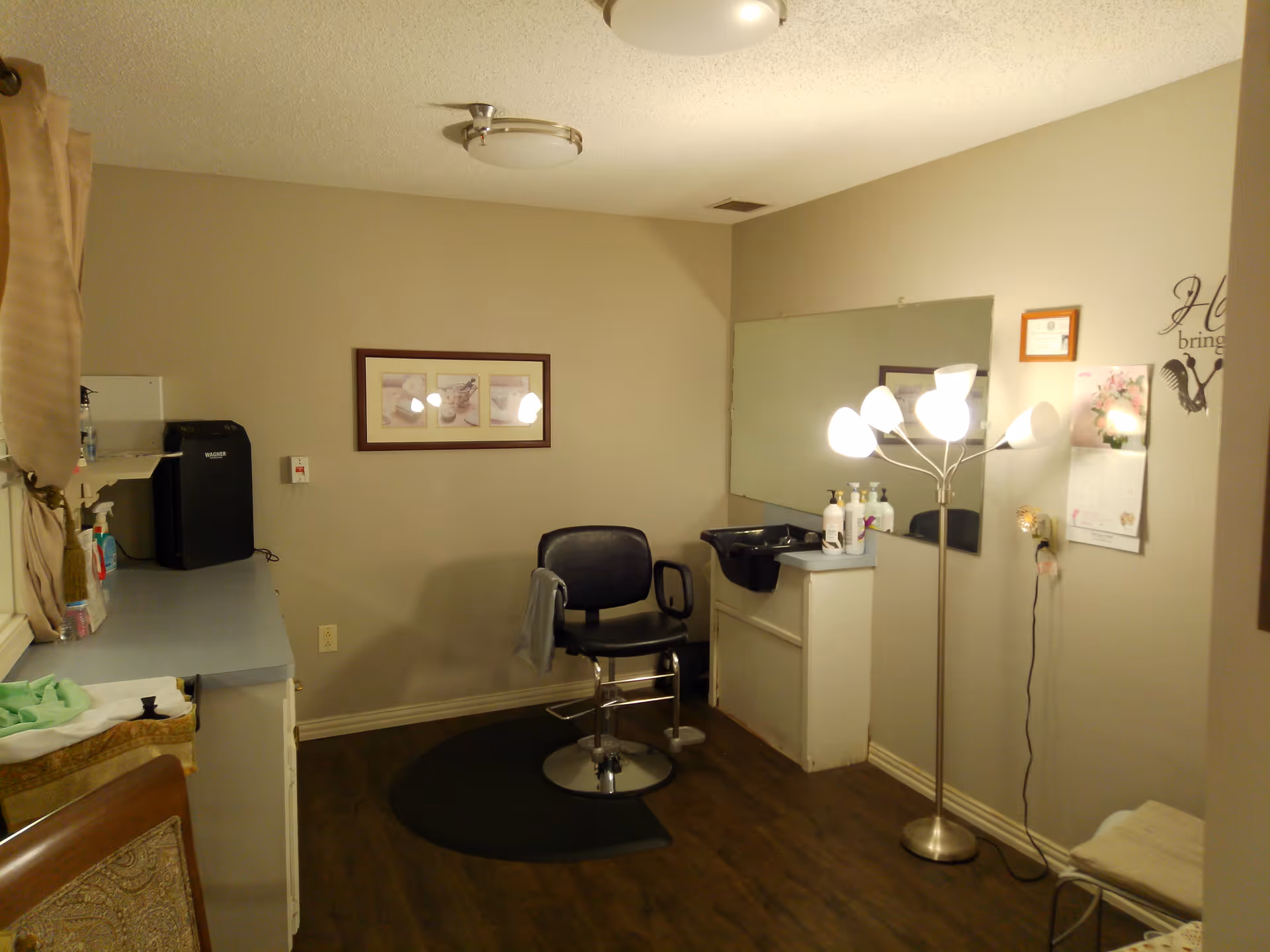 A small room with beige walls and wooden floor featuring a black salon chair on a black mat, a white counter with various items, a black sink, a tall floor lamp with multiple lights, a large wall mirror, framed pictures, and a calendar on the wall.