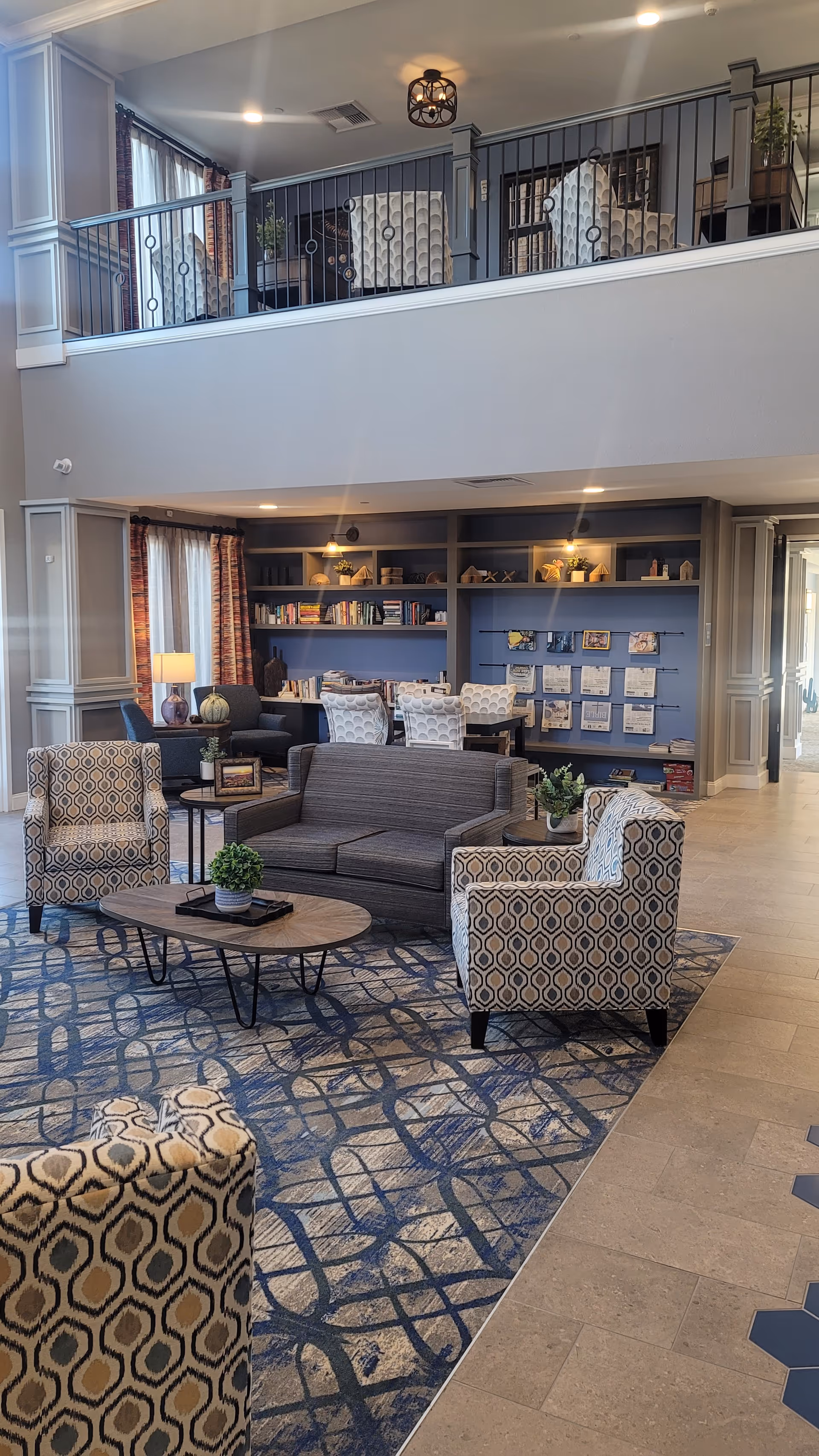 Spacious two-story community lounge with patterned armchairs, a sofa, coffee tables, bookshelves and a second-floor balcony seating area.