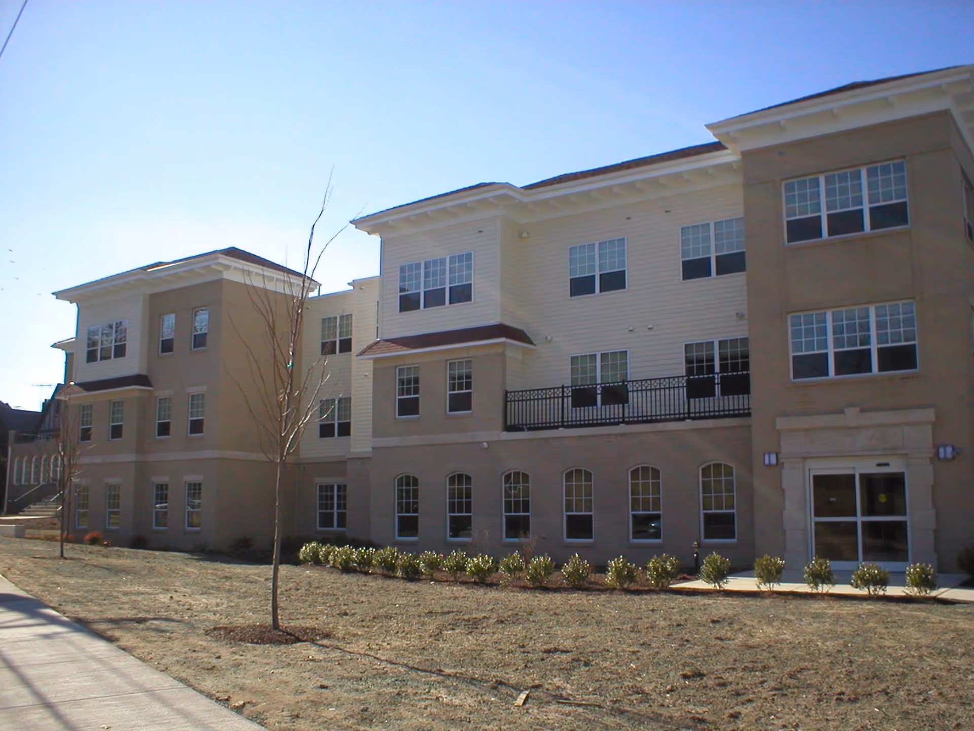 Exterior view of a three-story senior living facility building with multiple windows, a main entrance with glass doors, small trees, and shrubs planted in front under a clear blue sky.