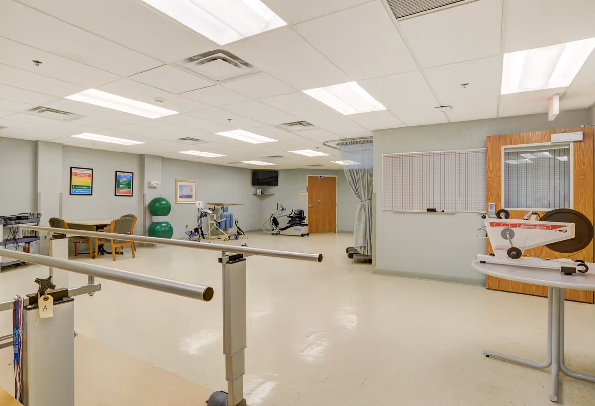 A spacious physical therapy room with parallel bars for walking exercises, exercise equipment including stationary bikes and therapy balls, a table with chairs, and a wall-mounted TV. The room has bright overhead lighting and a door with a window.