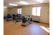 Small exercise room with several recumbent stationary bikes on a wood floor beneath fluorescent lights and windows.