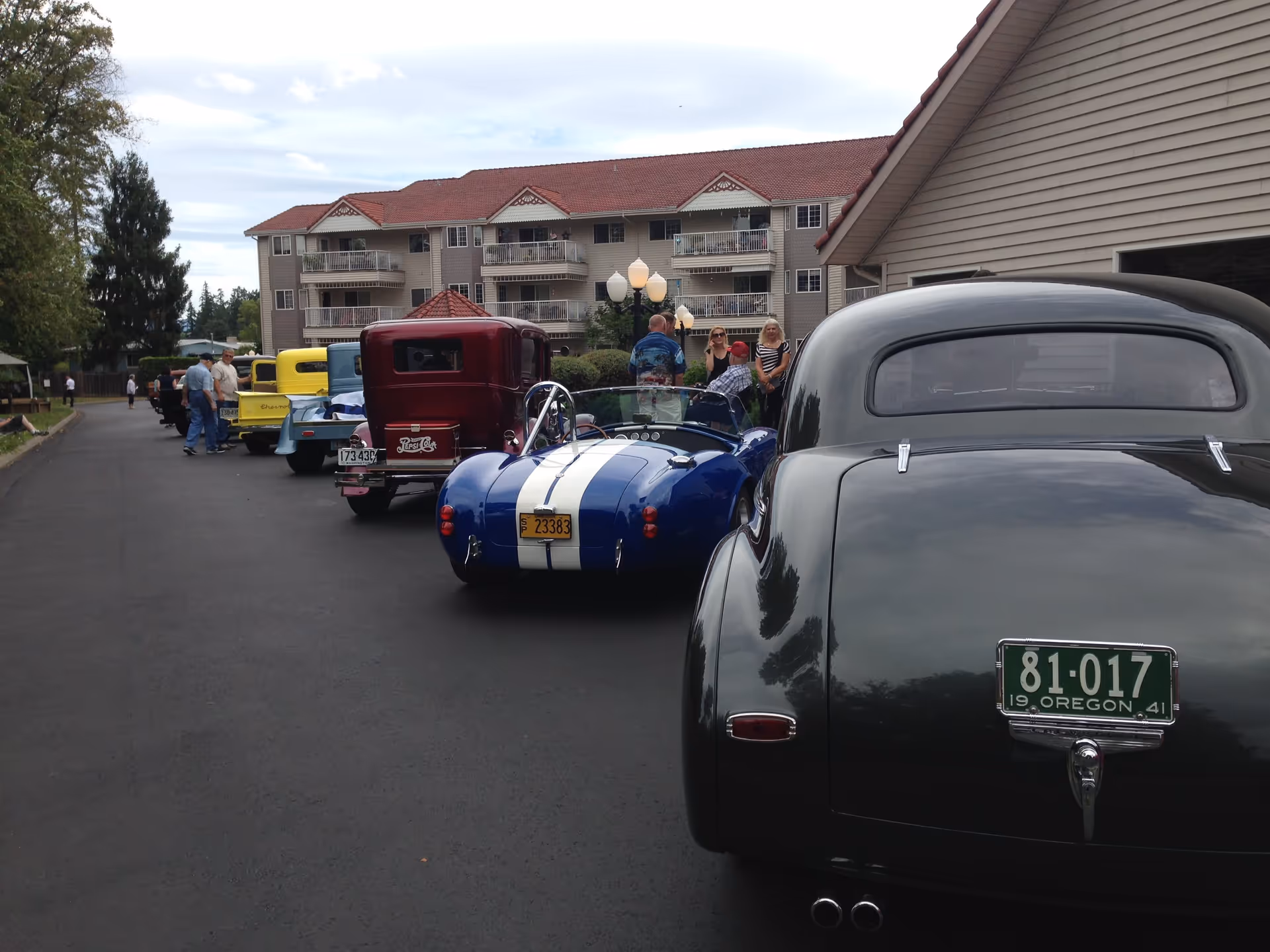 A lineup of vintage cars parked on a paved driveway outside a multi-story residential building with balconies. Several people are standing and conversing near the cars. The sky is partly cloudy and there are trees and street lamps along the driveway.