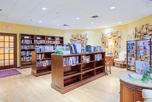 A bright and spacious library room with wooden bookshelves filled with books, a round table with magazines, comfortable seating including an armchair with a lamp, and large windows with patterned curtains. The room has light-colored walls and a wooden floor.