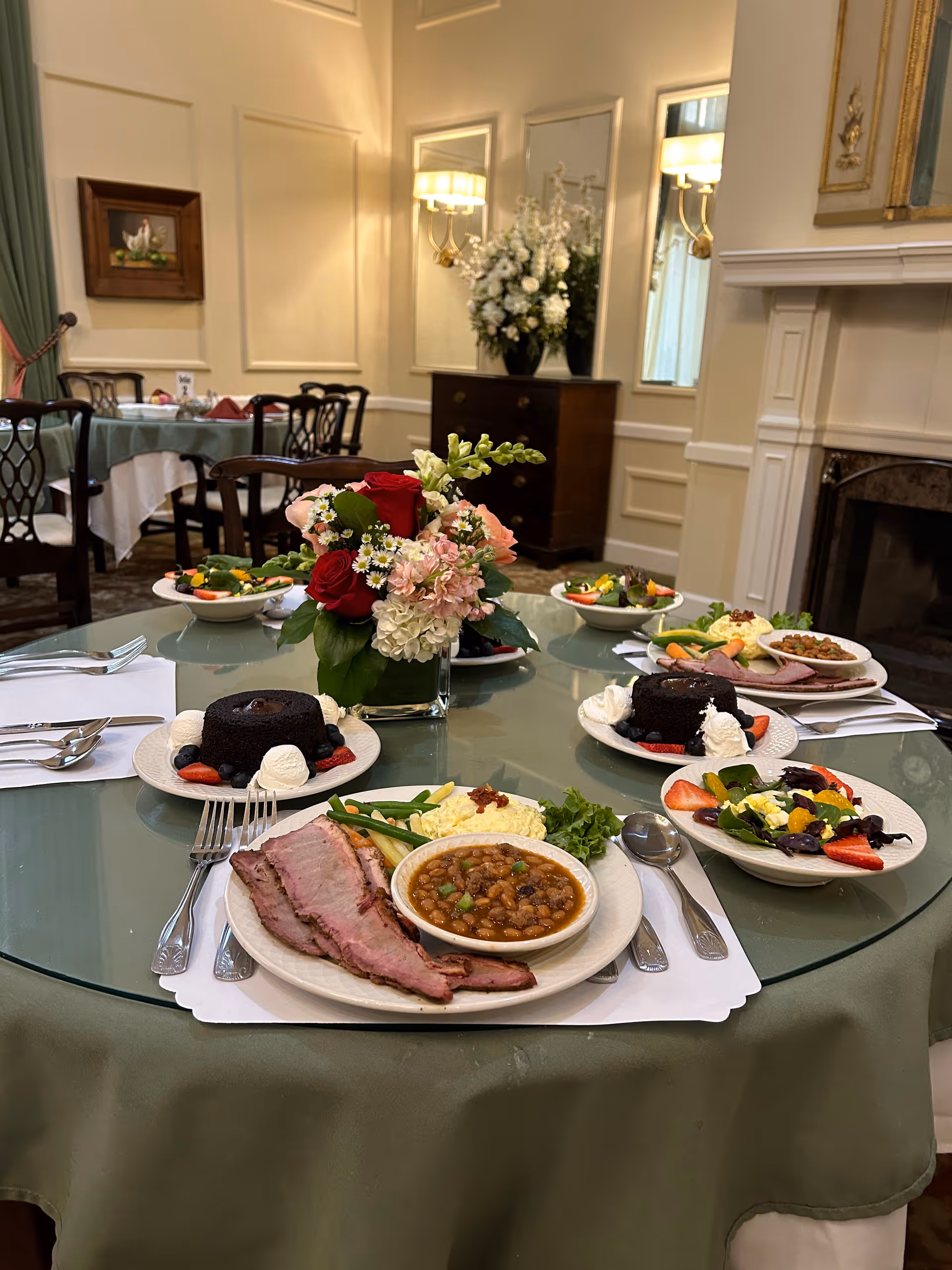 A dining table set with plates of food including sliced meat, baked beans, mashed potatoes, green beans, salad with fruit, and chocolate cake with whipped cream. The table has a green tablecloth and a floral centerpiece with red, pink, and white flowers. The background shows a classic dining room with wooden chairs, a sideboard with a large floral arrangement, wall sconces, and a fireplace.
