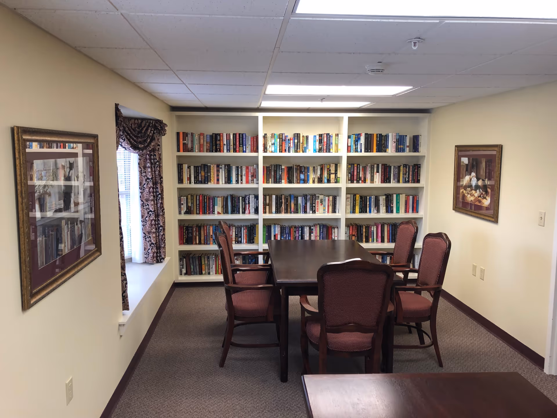 A quiet room with a large wooden table surrounded by six upholstered chairs. Behind the table is a white bookshelf filled with numerous books. The walls are light-colored and decorated with framed pictures. A window with patterned curtains is on the left side, allowing natural light into the room.
